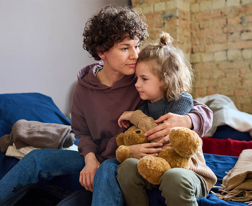 An adult sits on a bed holding a young child who is hugging a teddy bear, both looking thoughtful in a modest living space.