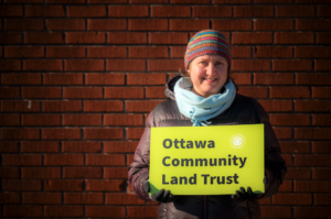 A person wearing a striped knit hat, light blue scarf, gloves, and a dark winter jacket stands in front of a red brick wall, smiling while holding a bright yellow sign that reads “Ottawa Community Land Trust.”