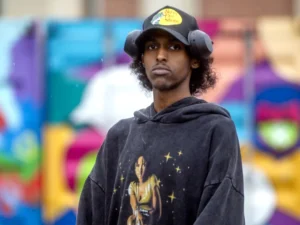 Alt text: Portrait of Idris Isse standing outdoors in front of a colorful blurred mural. He is wearing a black hoodie with a graphic print, a black baseball cap, and over-ear headphones resting around the sides of the cap. He has curly hair and a neutral expression while looking toward the camera.