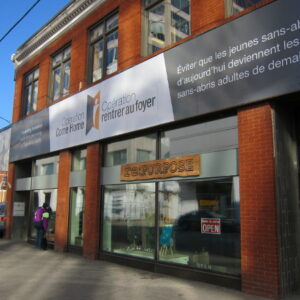 Alt text: Street-level view of a red brick building with large bilingual signage for “Operation Come Home / Opération rentrer au foyer,” a youth homelessness support organization. The sign includes French text about preventing homeless youth from becoming homeless adults. A storefront entrance with glass windows and an “OPEN” sign is visible at street level, with two people standing near the entrance on the sidewalk.
