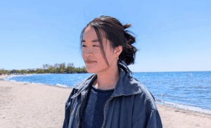 Photograph of a girl with black hair and a blue jacket standing in front of an ocean landscape